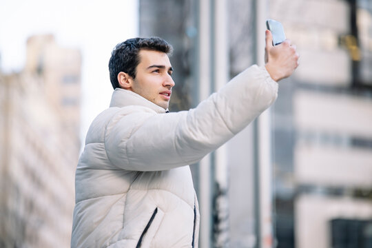 Side View Of Young Man In A White Puffer Jacket Takes A Selfie With His Smartphone On A City Street