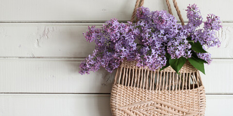 blooming branches of purple lilac in a woven summer bag hang on a wooden wall. Concept : no plastic.