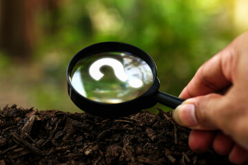Hands holding magnifying glass focused in question mark sign on the soil ground in green garden. Concept of quiz, many question arising on soil ground with natural environmental background.