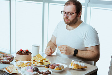 Man sitting in front of a table full of sweet food, eating pastry with cream. Fat man sugar addict, can't say no and stop eating, weight loss problem. Depressed man eats a lot of unhealthy meal.