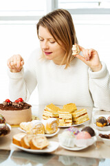 Woman eating pastry with cream, picking what to eat sitting beside table full of sweets. Diet is over, sugar addiction and problem with diabetes.