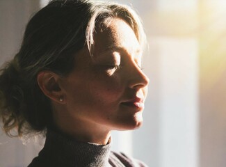 Serene Woman in Sunlit Room Practicing Mindfulness. A tranquil image woman with closed eyes, basking in sunlight, exuding peace and relaxation through breathwork or meditation.