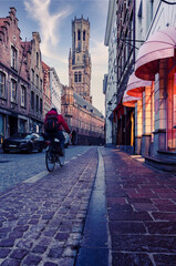 Cyclist riding past illuminated shop fronts on a cobbled street with the Belfry of Bruges in the background