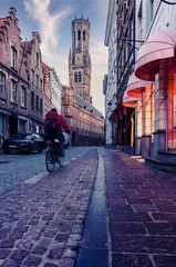 Tuinposter Cyclist riding past illuminated shop fronts on a cobbled street with the Belfry of Bruges in the background © ADDICTIVE STOCK CORE