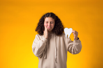 Child girl holding papercraft tooth and touching her tooth suffering fron toothache over yellow background. Dental health concept