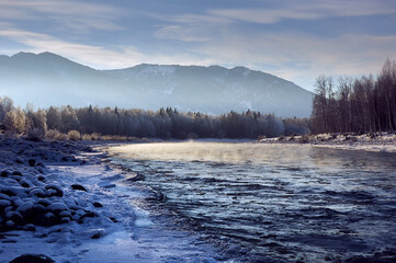 Snowcovered mountain landscape with frosty fog over a river and cloudy sky.