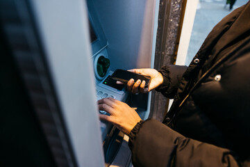 Close up of woman's hands using a smartphone with contactless technology at an ATM. Taking money...