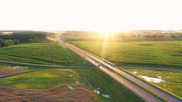 Vehicles fast driving on asphalt multi lane expressway. Intercity road passes through rural fields of growing corn and wheat. Cars drive on highway against sunshine rays at sunset. Aerial motion