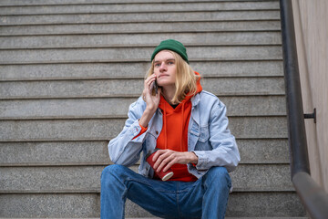 Calm hipster blond guy with coffee cup in hand sits on steps talks on smartphone in city street. Scandinavian man resting after long walk in downtown, sitting on stairs outdoors. 