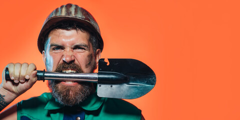 Angry bearded man in hard hat and uniform with shovel in teeth. Foreman, miner or builder in protective helmet. Mechanical or industrial worker in safety hardhat with spade. Copy space for advertise.