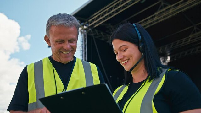 Production Team With Headsets And Clipboard Setting Up Outdoor Stage For Music Festival Or Concert