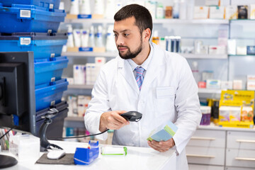 Naklejka na ściany i meble Male pharmacist in lab coat standing at counter in drugstore and using barcode scanner to sale drug package.