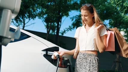 Young woman travel with EV electric car charging in green sustainable city outdoor garden in...