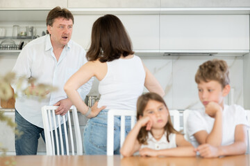 Brother and sister sitting at table in kitchen listening to quarrel of their mom and dad