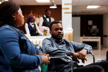 Young African American man in wheelchair in hotel lobby with able-bodied girlfriend, waiting for accessible room, handicapped guy traveling with mobility impairment. Accommodations for disabled - obrazy, fototapety, plakaty