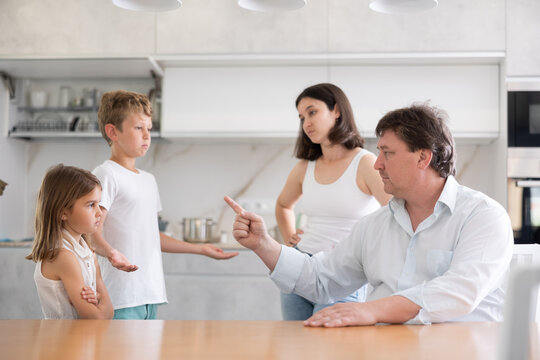 In kitchen, parents scold children for imprudent act. Dad explains to son and daughter about rules of safety behavior during independent walks. Children with guilty look stand in front of angry father