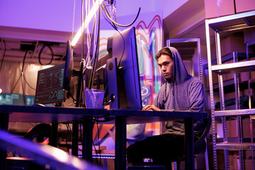 Asian hacker doing phishing attack while using illegal ransomware. Young internet criminal in hood installing malware for password cracking on victim computer in dark abandoned warehouse