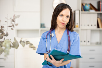 Waist-up portrait of woman medic standing in physician's office, holding paper folder.