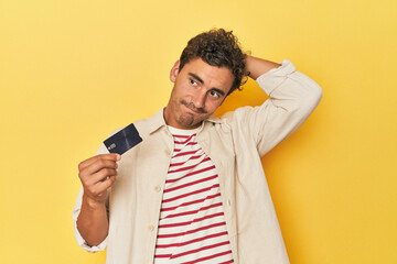 Man holding credit card on yellow touching back of head, thinking and making a choice. - obrazy, fototapety, plakaty