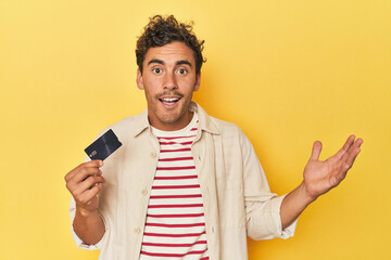Man holding credit card on yellow receiving a pleasant surprise, excited and raising hands.