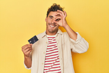 Man holding credit card on yellow excited keeping ok gesture on eye.