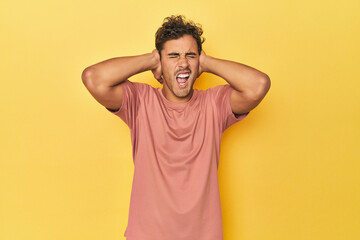 Young Latino man posing on yellow background covering ears with hands trying not to hear too loud sound.