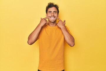 Young Latino man posing on yellow background smiles, pointing fingers at mouth.