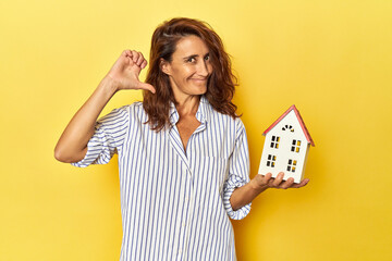 Middle aged woman holding a miniature house on yellow backdrop feels proud and self confident,...
