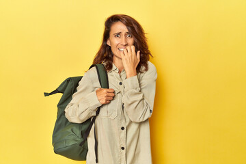 Middle aged woman ready for travel with backpack on yellow biting fingernails, nervous and very anxious.