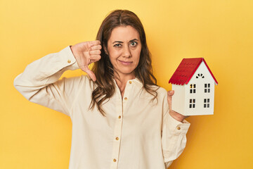 Holding miniature house, realty concept showing a dislike gesture, thumbs down. Disagreement...