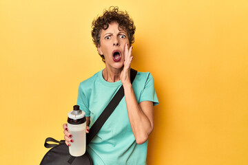 Sportswoman with gym bag & water on yellow studio shouting and holding palm near opened mouth.
