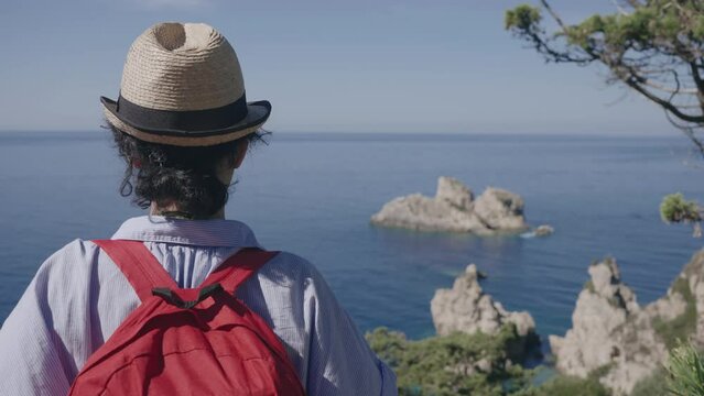 Tourist woman lwith straw hat and backpack ooking at amazing rocky coas landscape from Paleokastritsa monastery on Corfu island in Greece