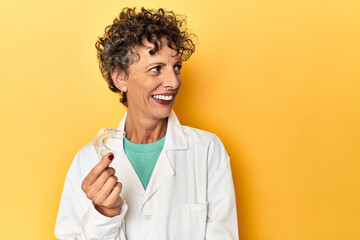 Doctor with invisible dental aligner on yellow looks aside smiling, cheerful and pleasant.