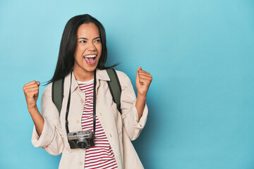 Filipina woman with camera and backpack on blue raising fist after a victory, winner concept.