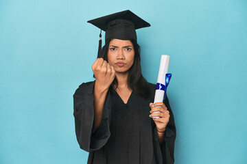 Filipina graduate with diploma on blue studio showing fist to camera, aggressive facial expression.
