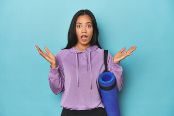 Filipina athlete with yoga mat on blue surprised and shocked.