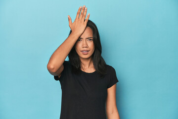 Filipina young woman on blue studio forgetting something, slapping forehead with palm and closing...