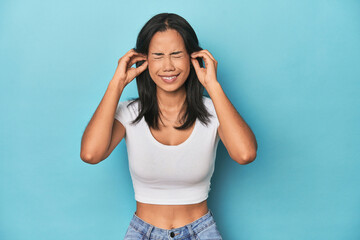 Filipina young woman on blue studio covering ears with hands.