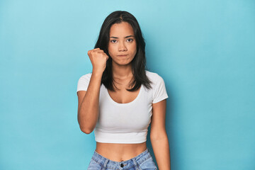 Filipina young woman on blue studio showing fist to camera, aggressive facial expression.