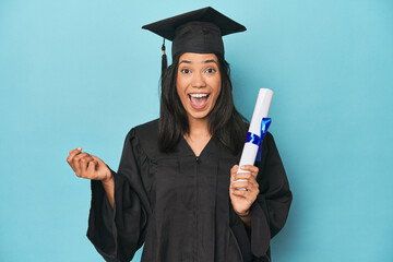 Celebrating Filipina graduate with diploma and cap on blue studio - obrazy, fototapety, plakaty