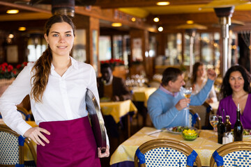 Portrait of confident smiling young waitress standing with serving tray in busy restaurant hall....