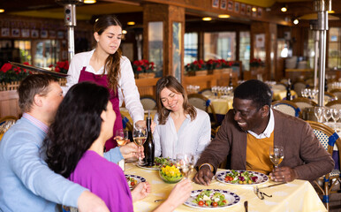 Young waitress serving bottle of wine to cheerful multiracial group of adults having friendly get-together in cozy restaurant..