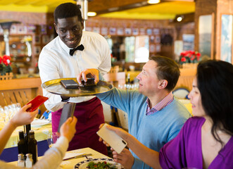 African-american man waiter standing with tray and taking away phones from group of people in...