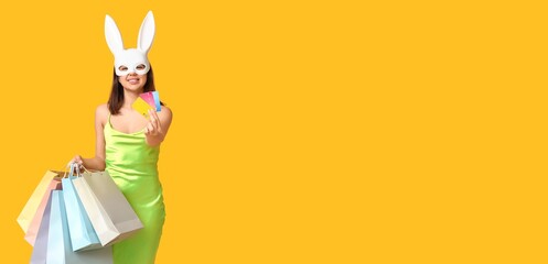 Beautiful young woman in bunny bask holding shopping bags and credit cards on yellow background...