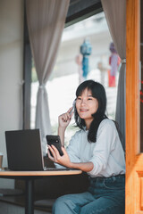 Obraz premium Business freelance concept, An Asian businesswoman sits thoughtfully in a cafe, holding a smartphone in one hand with a laptop open in front of her.