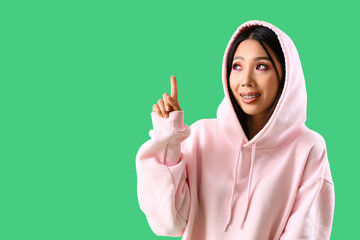 Young Asian woman in pink hoodie pointing at something on green background
