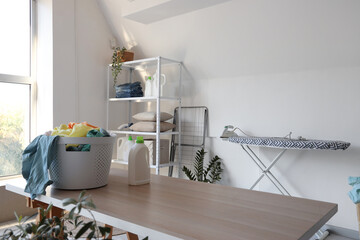 Interior of laundry room with clothes in basket, table, ironing board and shelving unit