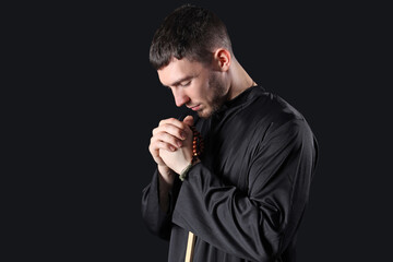 Young priest praying with beads on black background