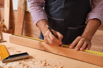 Mature carpenter measuring wooden plank at table in workshop, closeup