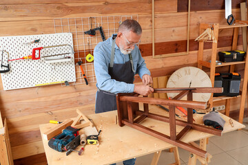Mature carpenter measuring wooden stool at table in shop